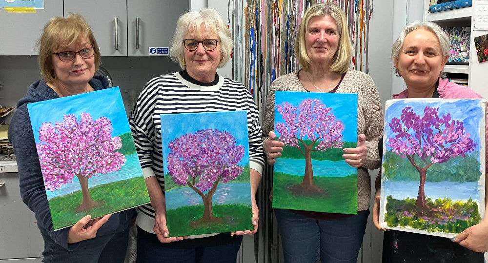 Four women holding paintings of trees with pink blossoms in a studio setting.