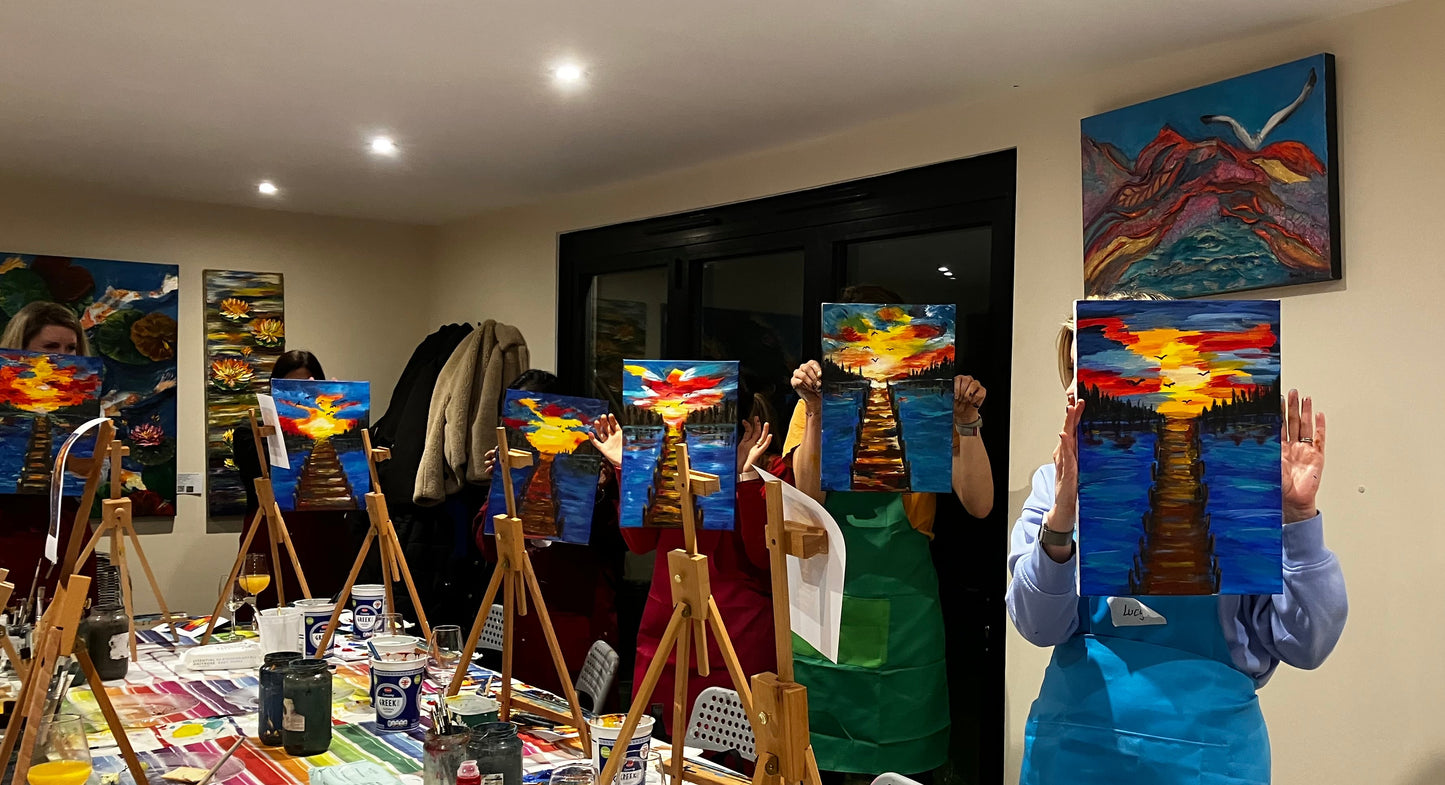 Art class with participants holding up their paintings in a room with easels and art supplies.