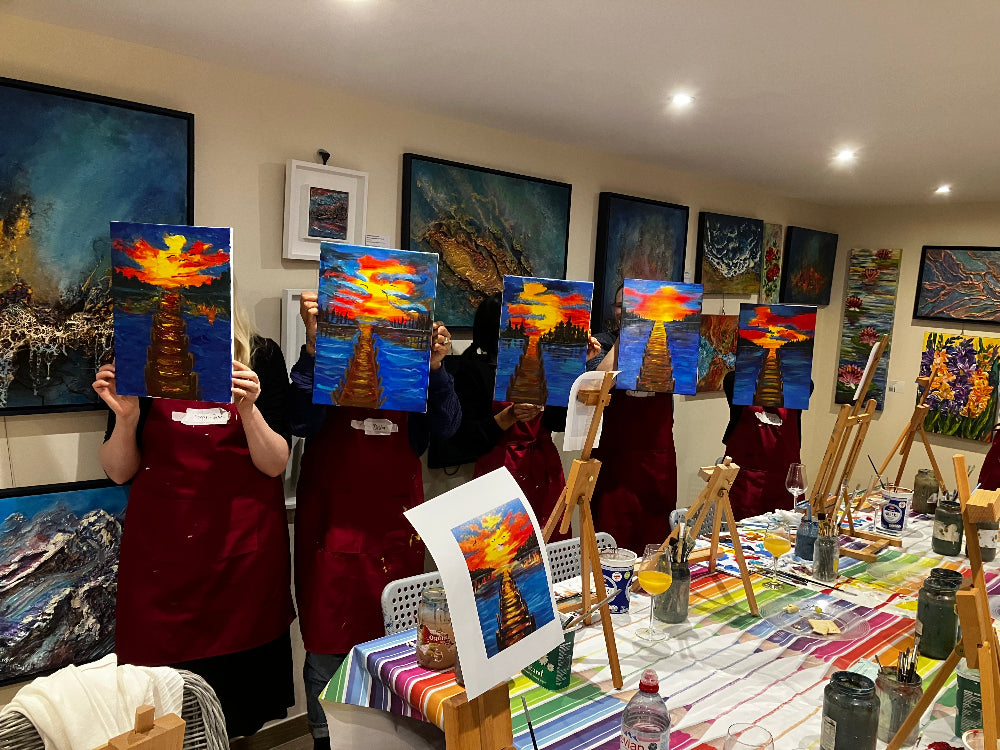 Group of people holding up paintings in a room with art supplies and easels.