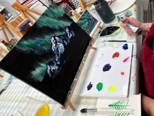 A person painting an aurora borealis landscape on a canvas at an art class or workshop, with a palette of mixed colors, paint brushes, and other painting supplies on a table.
