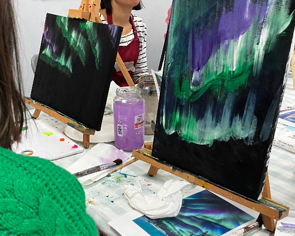 A group of people painting in a studio, with two easels displaying vibrant aurora-like artworks in green, purple, and black. One person in the foreground wears a bright green knit sweater, and other painting supplies and colorful wall art are visible.