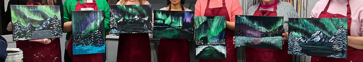 Seven women wearing red painterly aprons stand in an art studio holding small paintings of the green aurora borealis over snowy mountains. A paintbrush display hangs on the wall behind them, and painting supplies are visible on tables in the foreground.