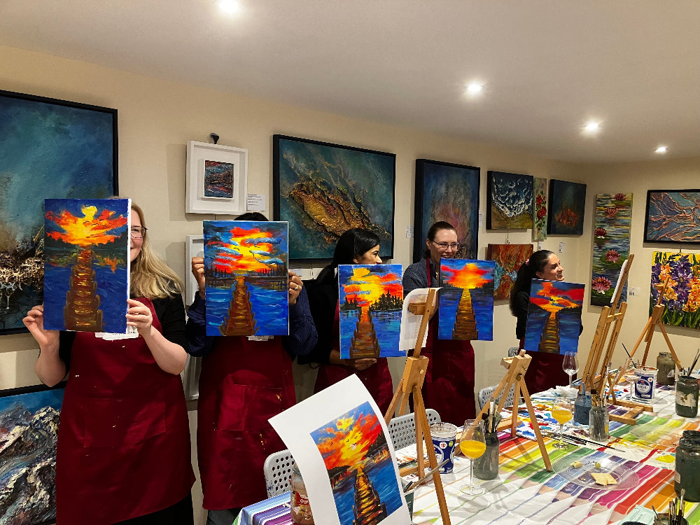Group of people holding up their paintings in a room with art supplies and other artworks on the walls.